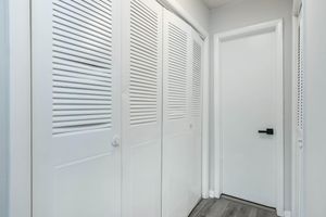Interior view of a hallway featuring white louvered closet doors on the left and a solid white door on the right. The walls are painted a light color, and the flooring is a light wood, creating a clean and modern aesthetic. Soft lighting enhances the space's brightness.