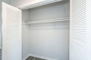 A spacious, empty closet with white louvered doors opened wide. The interior features a light gray wall and a single white shelf running along the back, with no items or clothing present. The flooring is a light wood look, adding to the clean and simple design of the space.