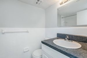 A clean and modern bathroom featuring a white tiled wall, a dark countertop with a sink and faucet, a large mirror above the sink, and a toilet. The space is well-lit with natural light and is organized with a towel bar installed on the wall.