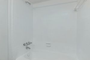 A clean and empty bathroom featuring a white tiled bathtub and shower area. The walls are smooth and white, with a chrome faucet and controls. There is a showerhead mounted on the wall, and a grab bar is installed for safety. The overall ambiance is bright and minimalistic.