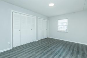 A bright, empty room with light gray walls and a smooth, dark gray laminate floor. There are two white closet doors with louvered panels on the left side, and a small window with blinds on the right. The ceiling features a round light fixture, creating a clean and modern atmosphere.