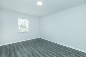 A bright, empty room with light gray walls and a window letting in natural light. The floor features dark wood-like laminate, and there's a ceiling light providing illumination. The room appears spacious and clean, ready for furnishing or personal touches.
