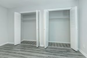 Two open closet doors with louvered panels, revealing empty interior spaces. The closets are set against a light gray wall, and the flooring is a light wood laminate. Natural light illuminates the area, creating a clean and spacious look.