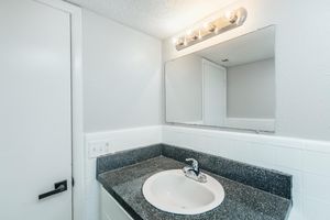A bathroom with a white sink and a dark countertop. Above the sink, there is a rectangular mirror and a light fixture with four bulbs. The walls are painted light gray, and a door is visible on the left side of the image. The overall look is modern and minimalist.