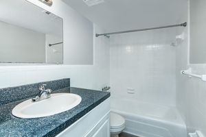 A clean and modern bathroom featuring a white tub with a shower, a large mirror above the sink, and a dark granite countertop. The walls and flooring are light-colored, creating a bright and spacious feel. Basic fixtures and a towel bar are also visible.