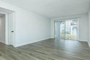 A spacious, empty room with light-colored walls and new, laminate flooring. Large sliding glass doors lead to an outdoor area, allowing natural light to fill the space. The room has a modern feel, with no furniture or decorations, and a neutral color palette.