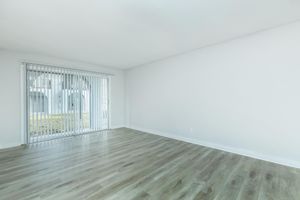 Spacious empty room with light wooden flooring and white walls. A large window with vertical blinds lets in natural light, creating a bright atmosphere. The room is devoid of furniture, offering a clean and minimalist aesthetic.
