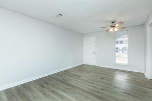 A clean, empty room with light grey walls and a ceiling fan. The floor features light wood-like laminate. A closed door is visible on the left, and a window with blinds allows natural light to enter. The room appears spacious and ready for furniture placement.