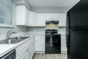 A modern kitchen featuring white cabinetry, a black stove and refrigerator, and double sinks. The space has gray walls, a light-colored countertop, and a window with blinds, providing a clean and minimalist aesthetic.