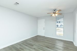 A bright, empty room with light gray walls and a wooden floor. A ceiling fan with five blades hangs from the ceiling, and there is a window covered with blinds on one side. The room includes a white door leading outside and a small amount of natural light coming through the window.