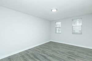 A minimalistic, empty room with light gray walls and a wooden floor. Two small windows with blinds let in natural light, and a flush ceiling light illuminates the space. The overall ambiance is bright and clean, emphasizing the room's openness and potential for decoration or use.