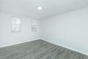 A minimalist, empty room with white walls and two small windows featuring blinds. The flooring is light-colored wood laminate. A circular ceiling light provides illumination. There are no furnishings or decorations, emphasizing the spacious feel of the room.