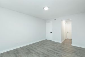 A bright, empty room with white walls and light gray laminate flooring. There's a ceiling light fixture, a closed door on the left, and an open door leading to another room on the right. The space is uncluttered, giving it a minimalist look.