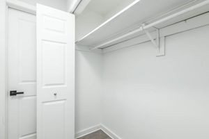 A minimalist, empty closet with white walls and shelving. The closet features a single door, partially open, revealing the interior space that is free of any items, conveying a clean and organized look. The floor is light-colored, enhancing the bright, airy feel of the space.