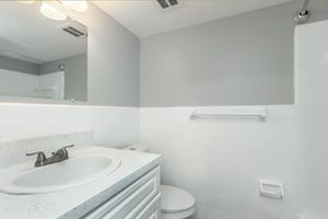 A clean and modern bathroom featuring a white tiled wall, a round sink with a faucet, a toilet, and a grab bar. The walls are painted in a light gray color, and there is a mirror above the sink. Soft lighting from overhead fixtures creates a bright ambiance.