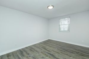 Empty room with white walls and a window featuring blinds. The floor has light brown wood-like laminate, and there is a ceiling light fixture. The space is well-lit and has a clean, minimalistic appearance.