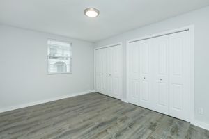 A bright, empty room with light gray walls and durable wood-like flooring. There are two white double doors on the right side, leading to closets, and a window with white blinds on the left, allowing natural light to fill the space. The ceiling features a simple light fixture.