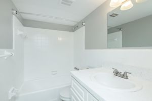 A clean and well-lit bathroom featuring a white bathtub with a shower rod, a white toilet, and a sink with a quartz countertop. The walls and tiles are predominantly white, and there is a mirror above the sink. Soft lighting from fixtures above illuminates the space.