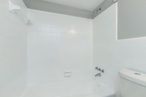 A clean and modern bathroom featuring a white tiled shower and bathtub. The shower area has a grab bar and a shower head, while the bathtub is empty. A white toilet is positioned beside the bathtub. The overall design is minimalistic with neutral colors.