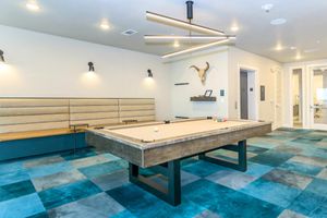 A modern game room featuring a pool table with a beige felt surface and colorful blue carpeting. There is a comfortable seating area with gray benches against the wall, a decorative wall-mounted bull skull, and contemporary lighting fixtures above. The room has a stylish, inviting atmosphere.