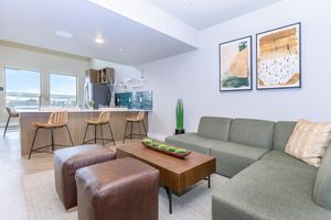 A modern living space featuring a green sofa and a wooden coffee table with decorative plants. To the left, there are two brown leather ottomans. The background includes a kitchen with bar stools and a stylish backsplash, along with two framed art pieces on the wall and large windows allowing natural light.