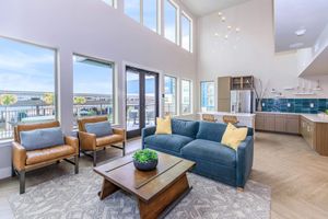 A spacious and modern living area featuring large windows with views, a blue velvet sofa with yellow pillows, two brown leather armchairs, and a wooden coffee table. The open kitchen area has sleek cabinets and a blue tiled backsplash, complementing the bright and airy atmosphere of the room.