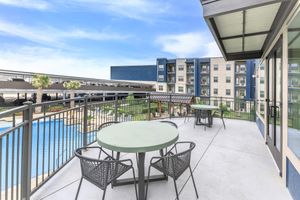 A spacious balcony with two round tables and chairs, overlooking a landscaped area and a swimming pool. In the background, modern apartment buildings can be seen under a clear blue sky. The setting conveys a relaxed and inviting atmosphere for outdoor gatherings.