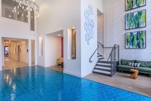 A modern interior lobby featuring a bright blue hexagonal tile floor, stylish green seating, and abstract wall art. The space is well-lit, with pendant lighting and a staircase leading to an upper level. Walls are painted in light colors, creating an inviting atmosphere.