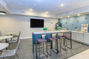 Modern lounge area featuring a large flat-screen TV mounted on the wall, a kitchenette with a decorative backsplash, a table with bar stools, and several small round tables with chairs. The space is brightly lit and has a welcoming, contemporary design, ideal for socializing or relaxing.