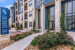 Exterior view of a modern apartment building named "The Current," showcasing a stone facade, large windows, and landscaped gardens. Flower beds with roses and small bushes are visible, along with a concrete pathway leading to the entrance. The building features a welcoming design with accessible pathways.
