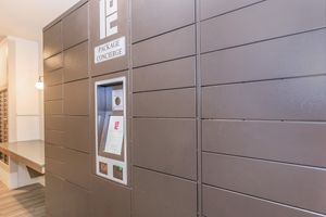 A modern package concierge locker system in a building lobby, featuring multiple horizontal compartments for parcel retrieval. The front panel displays a digital interface for user interaction, including a touchscreen and accessible slots for package securement. Ambient lighting enhances the contemporary design.