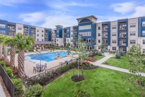 A vibrant apartment community featuring a landscaped courtyard with a swimming pool, lounge chairs, and palm trees. The buildings are modern with blue and beige exteriors, surrounded by neatly maintained gardens and walkways, providing a relaxing outdoor living space.