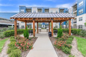 A well-maintained outdoor gazebo with a metal roof, surrounded by landscaped gardens and colorful flower beds. There are several tables and chairs underneath the gazebo, offering a space for relaxation. In the background, a modern apartment building can be seen.
