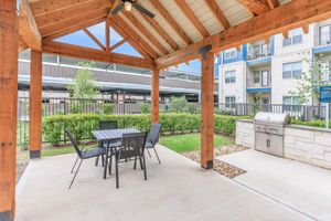 A covered outdoor seating area featuring a table with four chairs, a built-in grill, and landscaped greenery. The structure has wooden beams and a shingled roof, providing a shaded space for relaxation or gatherings in a modern residential setting.