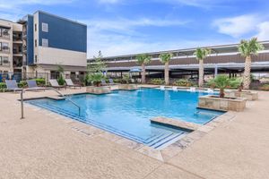 A clear blue swimming pool surrounded by lounge chairs and palm trees, with a modern apartment building in the background. The area features a landscaped garden and shaded parking spaces nearby. The sky is bright and clear, suggesting a sunny day.