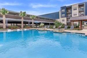 A sparkling swimming pool surrounded by palm trees and lounge chairs, with a modern apartment building in the background. The pool area features open spaces for relaxation and is well-maintained, creating a tranquil atmosphere for residents and guests.