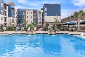 A vibrant outdoor swimming pool surrounded by lounge chairs and tropical plants. Modern apartment buildings are visible in the background, with a blue sky overhead. The setting exudes a relaxing and inviting atmosphere, perfect for leisure and enjoyment.