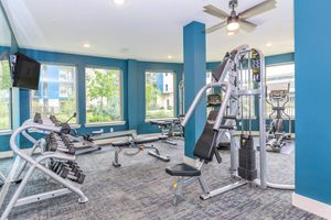 A well-lit gym area featuring various exercise equipment, including weight benches, dumbbells, and a multi-station weight machine. Large windows allow natural light to fill the space, and modern decor with blue accent walls creates a vibrant atmosphere.