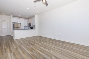 A spacious, modern apartment interior featuring light-colored walls and wood-like flooring. The room includes a ceiling fan, large windows, and an open layout leading to a kitchen area with stainless steel appliances. The kitchen is partially visible in the background, showcasing a clean and contemporary design.
