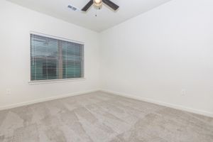 Empty room with light-colored walls and carpet. There is a large window with blinds, allowing natural light to enter. A ceiling fan is visible, and the space appears clean and well-maintained, with no furniture or decorations present. Ideal for personal customization.