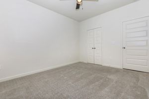 A minimalistic room featuring light gray walls and a light gray carpet. There is a ceiling fan with three blades and two white-paneled doors on the right side, leading to an adjacent space. The room is empty, creating a spacious and uncluttered atmosphere.