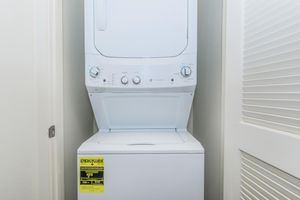 A compact white washer and dryer unit stacked together in a small laundry area. The dryer features control knobs on the front and an energy guide label indicating usage information. The background includes a light-colored wall and a partially open closet door.
