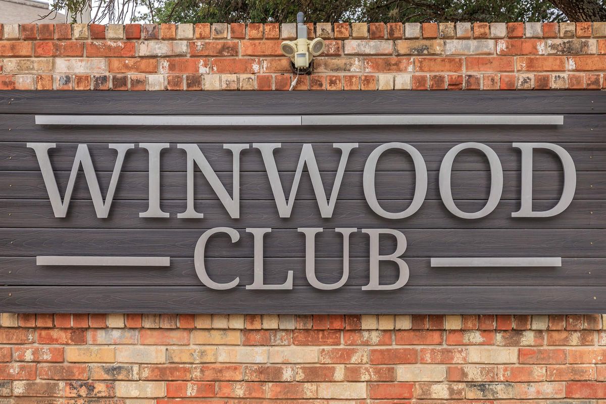 Sign for Winwood Club featuring large, bold letters on a wooden panel with a brick background. The lettering is in a modern font, prominently displaying the words "WINWOOD CLUB."
