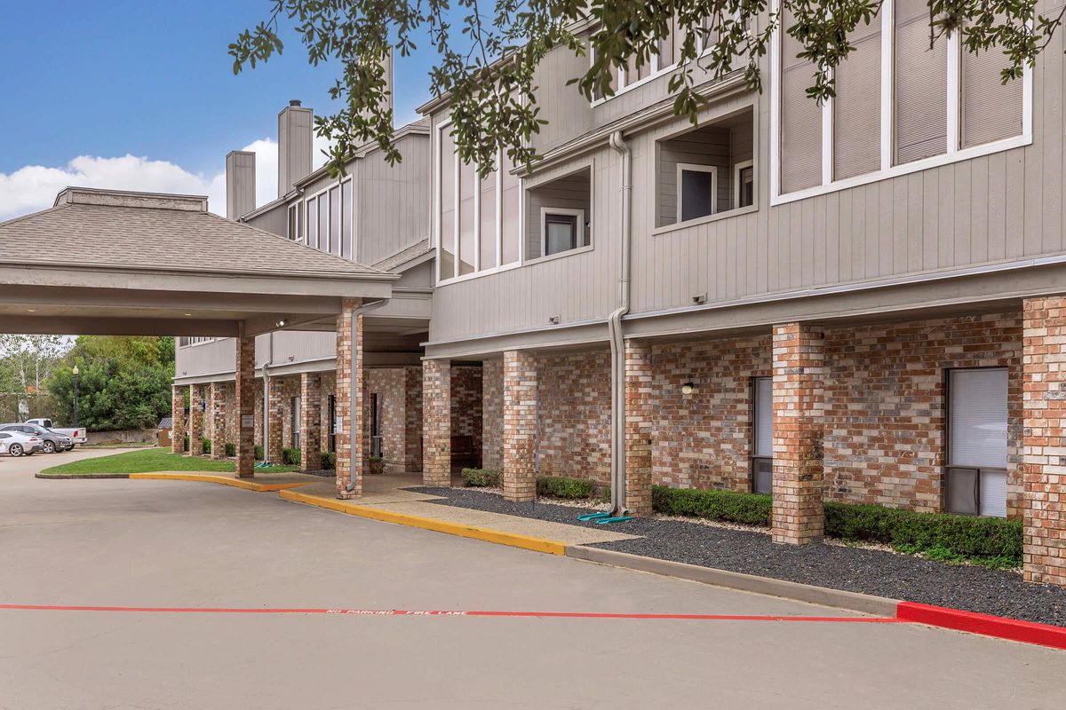 Exterior view of a multi-story building featuring a covered entrance and brick columns. The façade showcases a combination of gray siding and brickwork. Lush landscaping with low shrubs lines the entrance, and a clear blue sky is visible in the background.