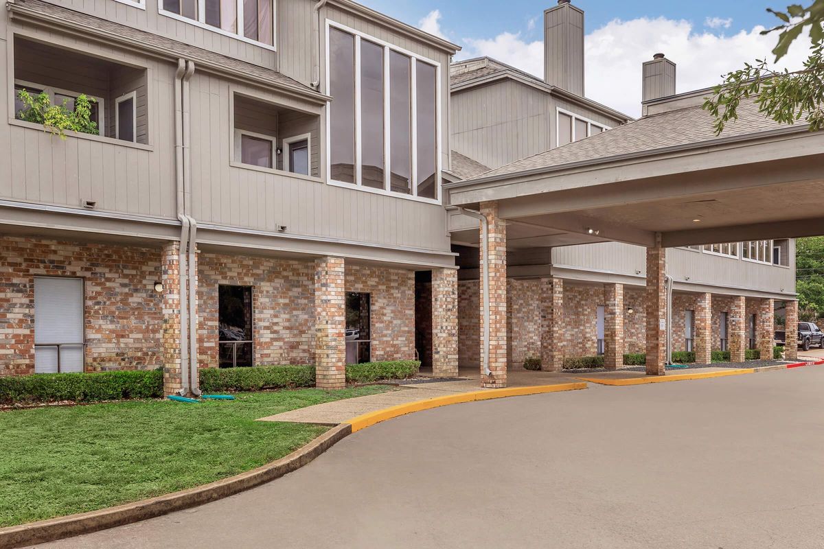 A view of a multi-story residential building with brick accents and large windows. The entrance features a covered area supported by columns, with a landscaped lawn and a curved driveway leading up to it. The sky is partly cloudy, adding a serene background to the scene.