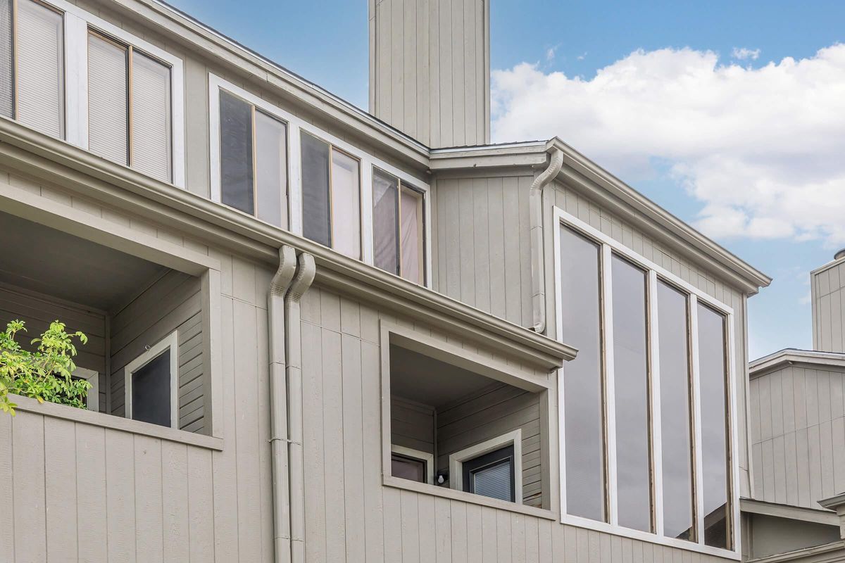 A modern, multi-story building with a light gray exterior. The structure features large windows, including a prominent section with floor-to-ceiling glass. The architecture includes a chimney and various balcony designs. The sky above is partly cloudy, creating a tranquil atmosphere.