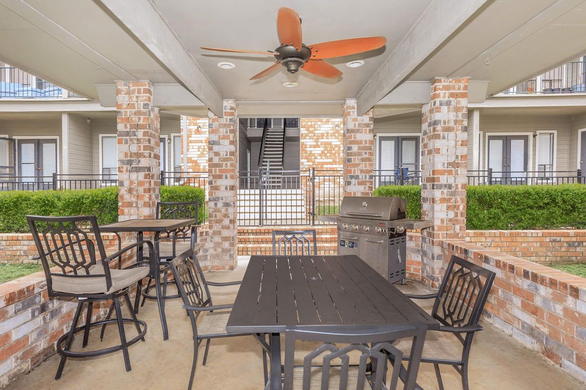 Outdoor seating area featuring a black metal table and chairs, a ceiling fan, and a barbecue grill under a covered patio. Surrounding the space are brick columns and neatly trimmed greenery, with staircases leading up to upper-level apartments in the background.