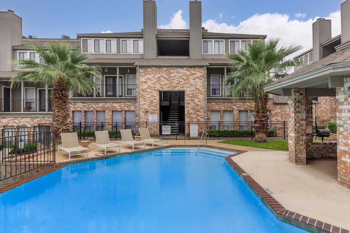 A clear blue swimming pool surrounded by lounge chairs, set against a backdrop of brick and modern apartment buildings. Lush palm trees add greenery, and the sky is partly cloudy. An inviting outdoor space for relaxation and leisure.