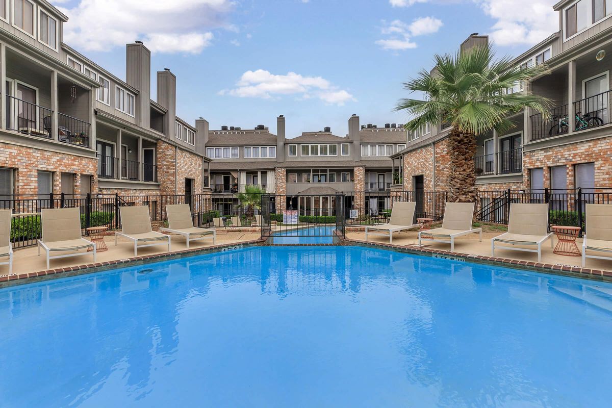 A clear blue swimming pool surrounded by lounge chairs, with multiple apartment buildings in the background featuring brick and stucco exteriors. Lush greenery and a palm tree add to the outdoor ambiance, set against a bright blue sky with scattered clouds.