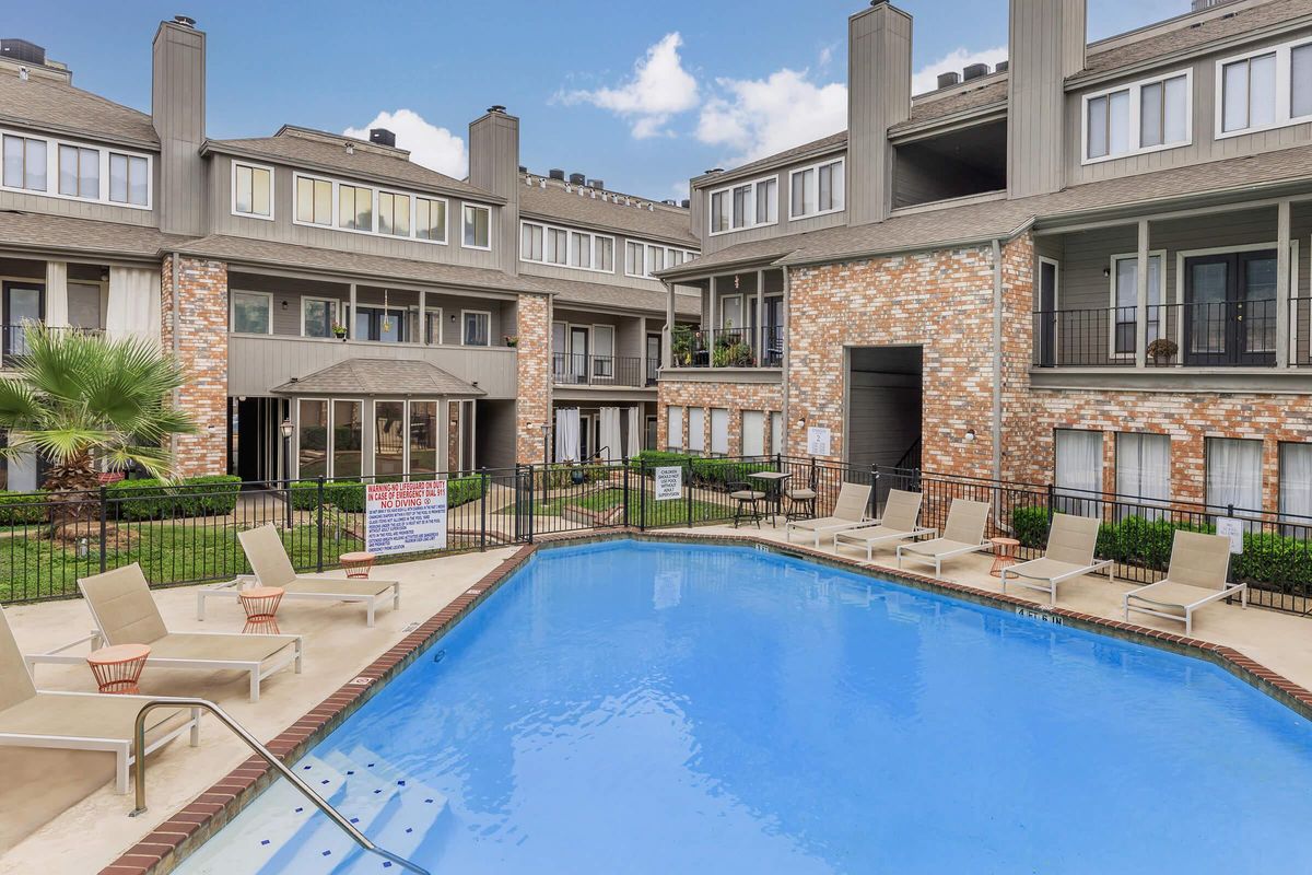 A tranquil swimming pool surrounded by a patio area with lounge chairs, located in a landscaped courtyard area of an apartment community. The buildings feature a mix of brick and siding, with palm trees and green grass enhancing the inviting atmosphere. Bright blue sky with fluffy clouds above.